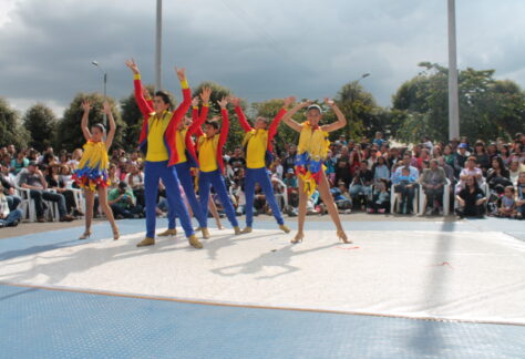 Presentación de un grupo de danza que se observa en el centro vestidos con los colores de la bandera de Colombia. En el fondo se observa un público amplio observando la presentación.
