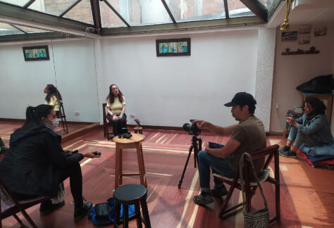 Cuatro personas se encuentran reunidas en un salón con piso de madera y un espejo en una pared. Dos personas se preparan con cámara y grabadora para realizar la entrevista a una mujer.