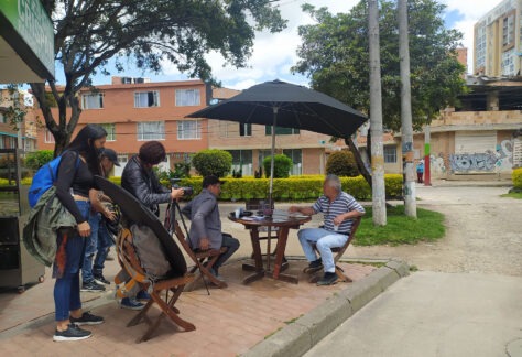 En una mesa con una sombrilla ubicada al aire libre en un andén, se reúnen una persona mayor con cuatro jóvenes que se preparan para grabar una entrevista.