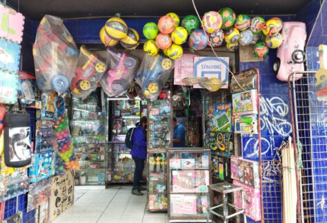 Mujer comprando en una juguetería. Se observan colgados sobre las vitrinas de cristal balones, pequeños carros plásticos, pelotas de colores y cestas.