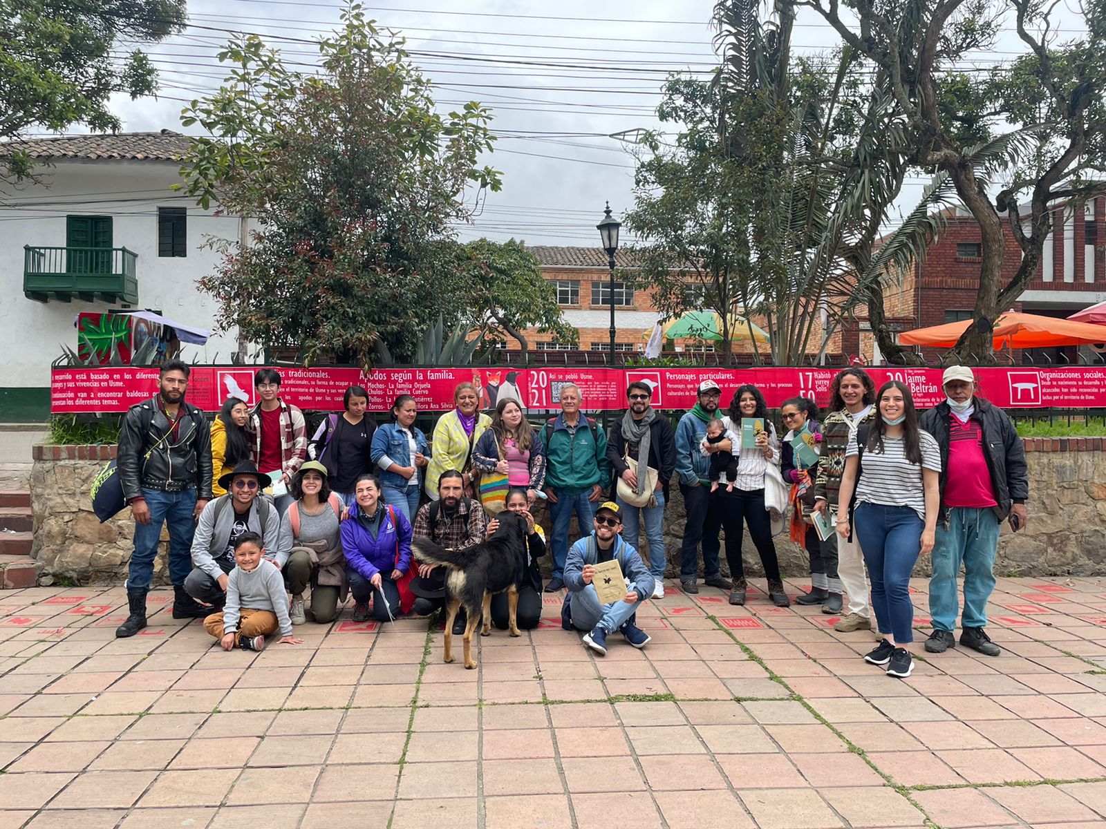 Grupo de personas posan en plaza pública para fotografía grupal. Se observa un perro en el centro y una tela roja detrás con información sobre el patrimonio de Usme.