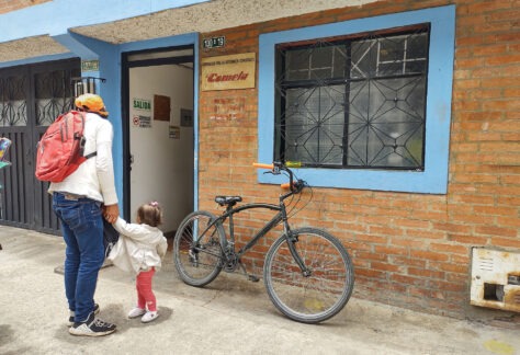 Hombre y niña pequeña tomados de las manos cerca a la puerta de un establecimiento denominado Cometa con una ventana de marco azul y bicicleta negra con manijas anaranjadas descansada sobre la pared de ladrillos.