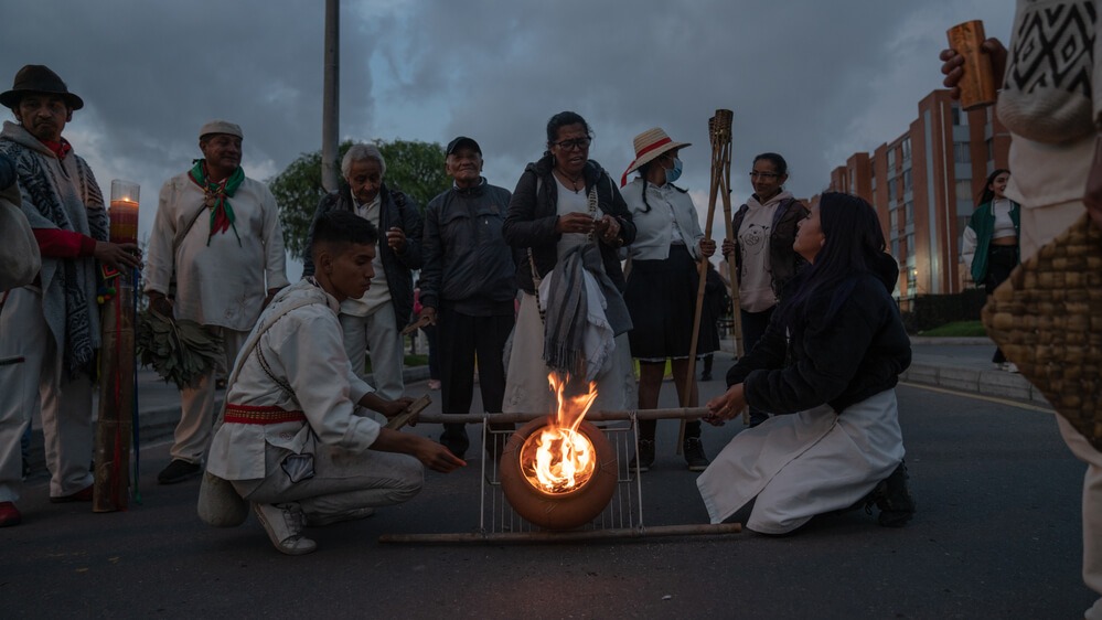 Autoridades hombre y mujer cargando una olla con fuego en las calles, alrededor personas del Cabildo Indígena Muisca de Bosa acompañando.
