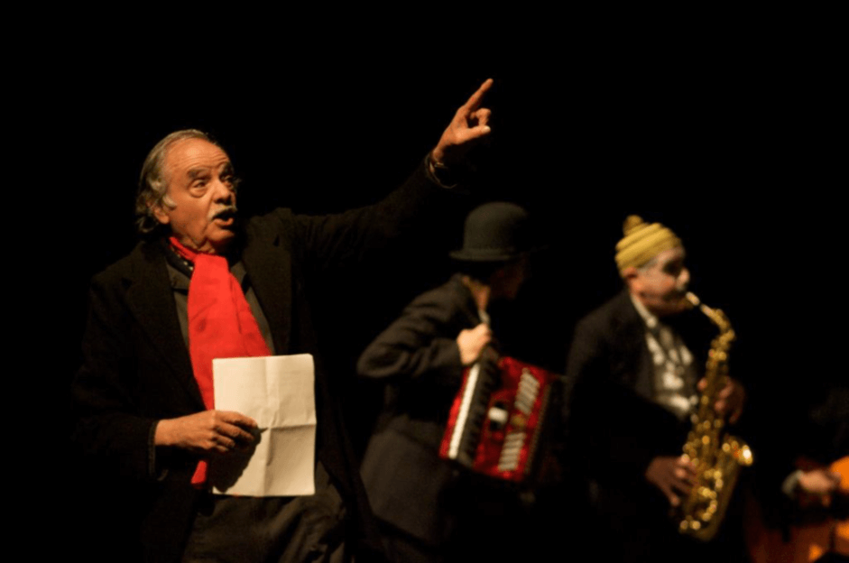 tres actores en un escenario teatral, uno de ellos es Santiago García, fundador del Teatro La Candelaria, quien aparece sosteniendo una hoja de papel con la mano derecha y apuntando al cielo con la izquierda