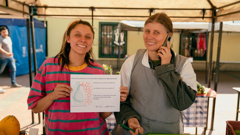 Dos mujeres sonriendo posan junto a un dibujo en la en el mercado campesino Agrocomunal en la Plaza Fundacional
