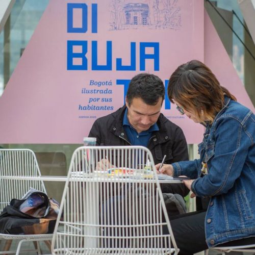 Dúo de participantes trabajando en sus ilustraciones frente al cartel de la Dibujatón.
