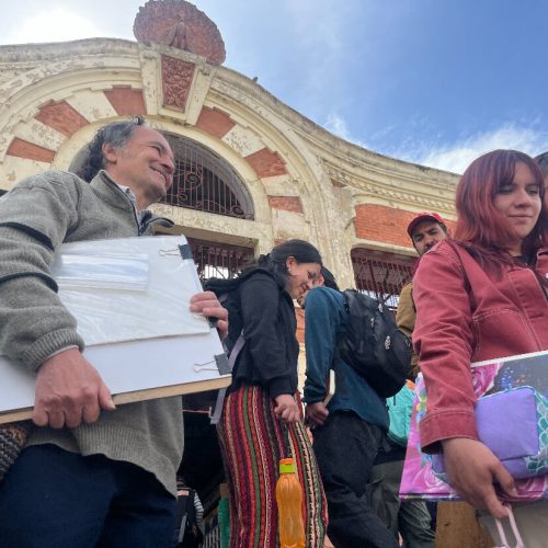 Personas participantes sostienen carpetas y dibujos frente a la fachada de la Plaza de Mercado Las Cruces durante el Premio Dibujatón 2025.