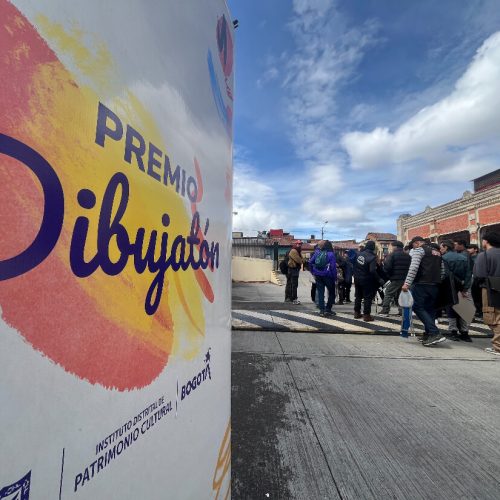 Pendón del Premio Dibujatón 2025 instalado en la Plaza de Mercado Las Cruces, con asistentes caminando alrededor.