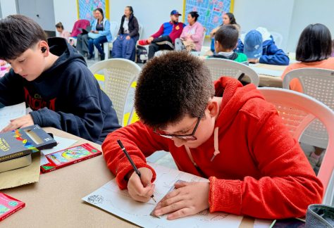 un niño traza con lápiz y regla el diseño de un vagón del Metro de Bogotá, al fondo, más participantes y acompañantes comparten el espacio de creación.