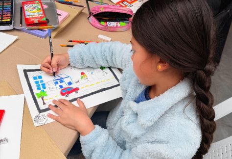una niña con trenza colorea una ilustración que incluye edificios, un carro y elementos de ciudad