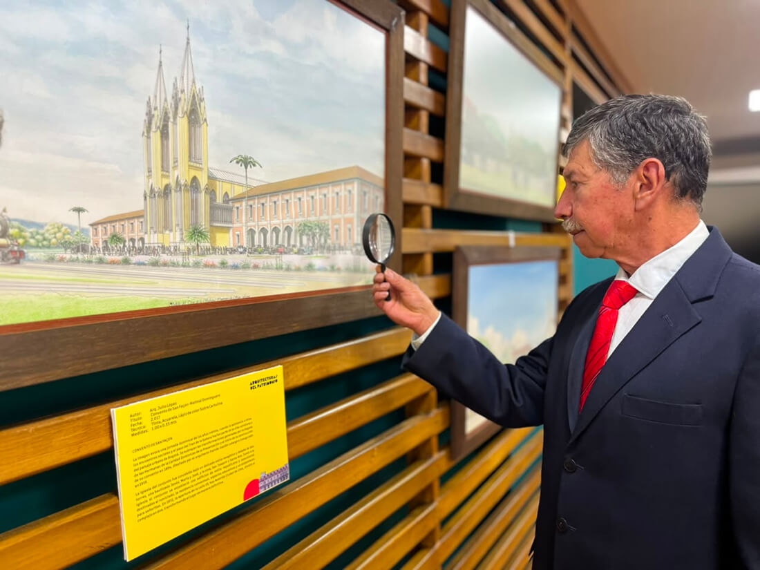 Hombre de traje examinando con una lupa una ilustración histórica enmarcada expuesta sobre un panel de exhibición.