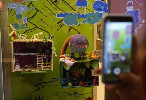 Primer plano de una mano sosteniendo un teléfono celular para tomarle una fotografía a los coloridos elementos internos y maquetas de papel expuestas en la muestra artística.