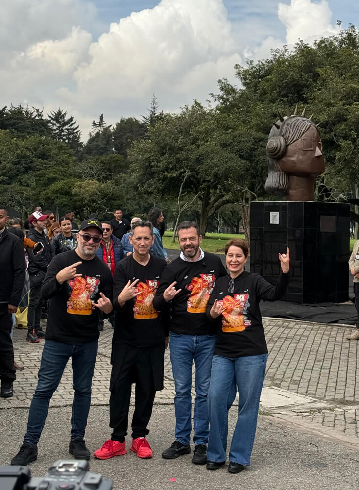 Grupo de personas posando sonrientes y haciendo el gesto rockero de la mano frente a la escultura de bronce "La Rockera" y su pedestal en un área verde del Parque Metropolitano Simón Bolívar.