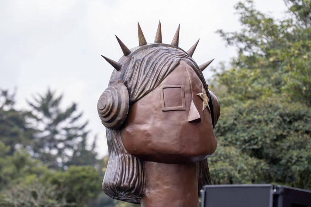 Primer plano de busto en bronce de la escultura "La Rockera" en el Parque Simón Bolívar. La obra representa una figura con audífonos intervenidos con taches en punta, una estrella tallada sobre uno de sus ojos y cabello estilizado.