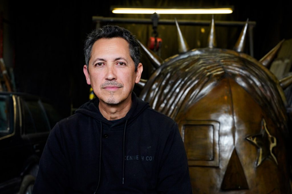 Retrato en primer plano del artista bogotano Maquiamelo en su taller, vistiendo una buzo oscuro y posando frente a la escultura en proceso de "La Rockera".