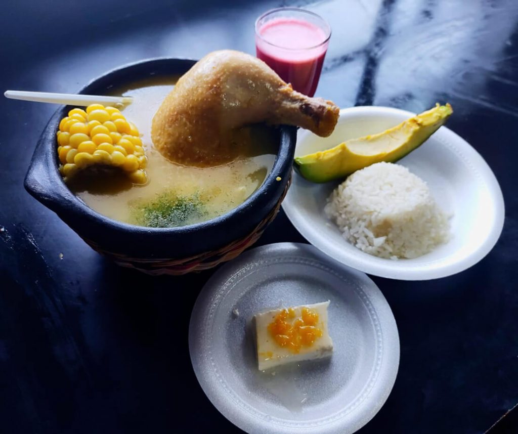 Plato de sopa tradicional acompañado de mazorca, una taza de chicha u otra bebida artesanal y una porción de amasijo en un plato.