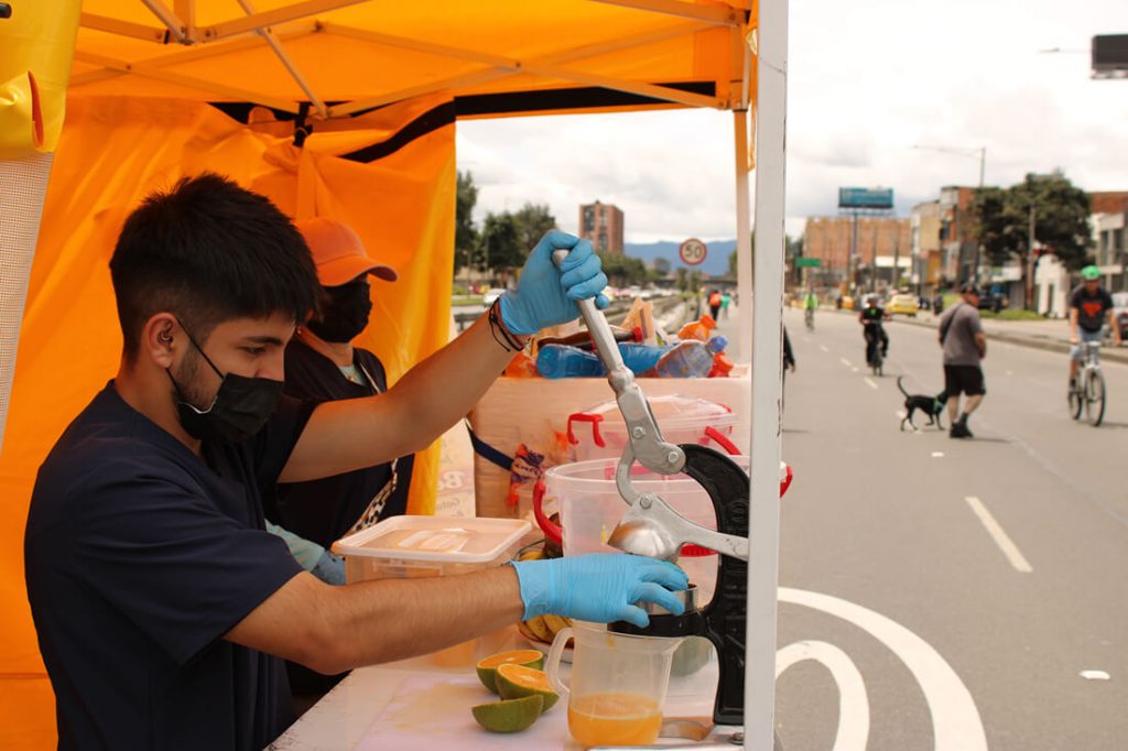 Hombre joven con tapabocas preparando un jugo de naranja durante la ciclovía, en una carpa amarilla.