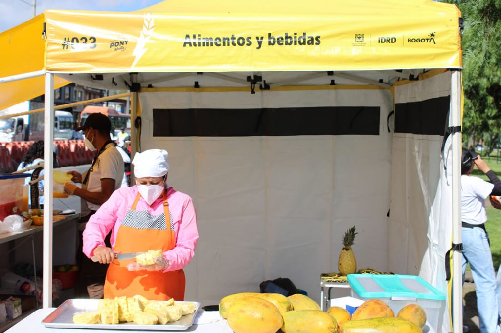 Puesto ambulante en la ciclovía de frutas y bebidas bajo un toldo amarillo con el letrero "Alimentos y bebidas".