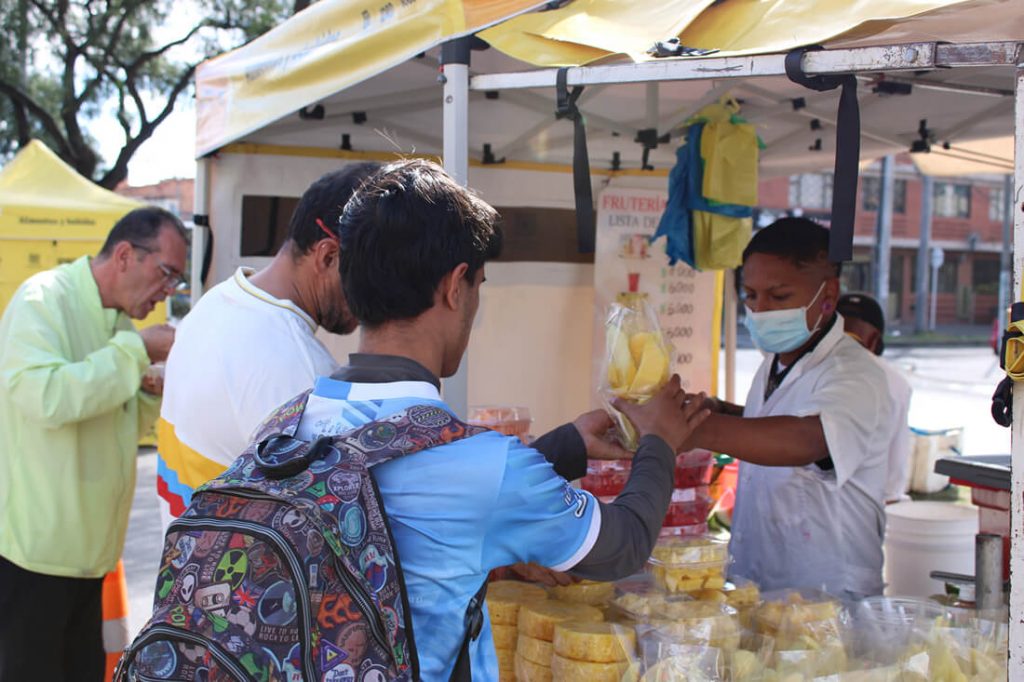 Un joven recibe una fruta en un plato en un puesto callejero con toldo blanco, en presencia de un comerciante con tapabocas.