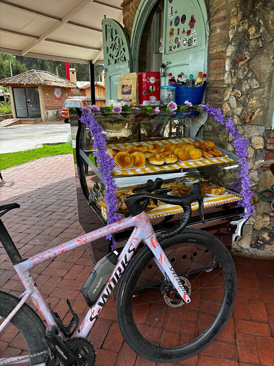 Una bicicleta de ruta parqueada frente a un puesto de alimentos tradicionales con panes, amasijos y refrescos exhibidos.