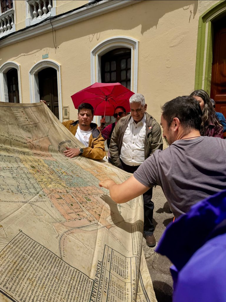 Un grupo de personas examina un mapa histórico desplegado al aire libre frente a una edificación colonial.