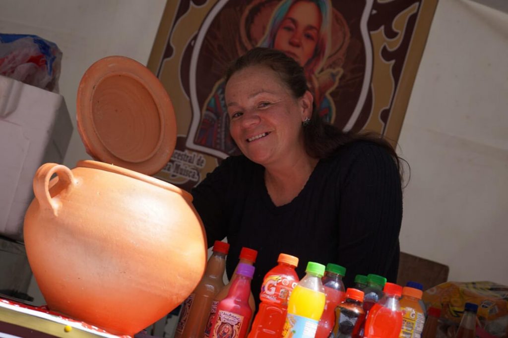 Una mujer sonriente vestida con ropa oscura sostiene un recipiente junto a una mesa cubierta con varias botellas de bebidas artesanales e industriales en recipientes de barro y plástico.