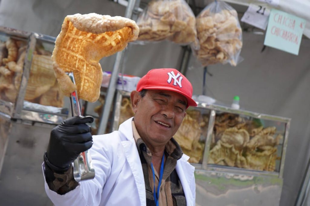Un hombre con gorra roja sostiene y muestra una pieza de comida frita dorada frente a su puesto de venta.