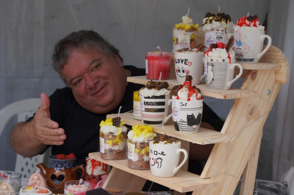 Hombre sonriente de pie junto a una mesa exhibidora cargada de pocillos, jarras decoradas y botellas de bebidas artesanales.