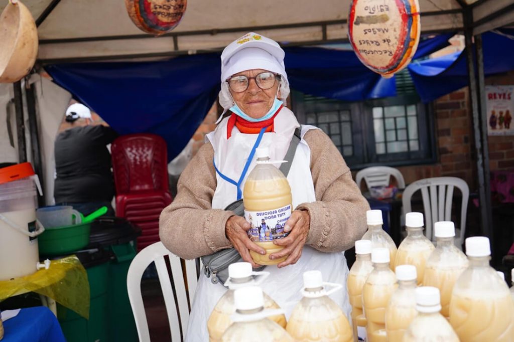 Persona mayor con delantal y gorra sosteniendo una botella de bebida artesanal frente a su puesto de venta.